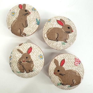 May include: Four round fabric covered knobs with a brown bunny rabbit design on a cream background. The bunnies have pink ears and are surrounded by small flowers and leaves.