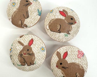 May include: Four round fabric covered knobs with a brown bunny rabbit design on a cream background. The bunnies have pink ears and are surrounded by small flowers and leaves.