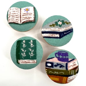 May include: Four round, green fabric-covered coasters with different book-themed designs. One coaster features an open book with a flower on the page, another has a stack of books with a mug on top, another has a single book with a floral design, and the last has a single book with a simple design.