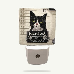May include: A novelty night light featuring a black and white cat mugshot design. The cat holds a sign that reads "Wanted Cat Burglary". The background has height markings in feet and inches.