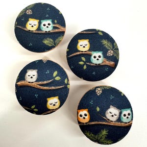 May include: Four round wooden knobs with a dark blue fabric covering. The fabric has a repeating pattern of owls on branches with pine needles and stars. The owls are yellow, orange, white, and blue.