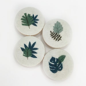Puede incluir: Cuatro posavasos redondos de tela con diferentes diseños de hojas verdes. Los posavasos son blancos con una textura sutil.