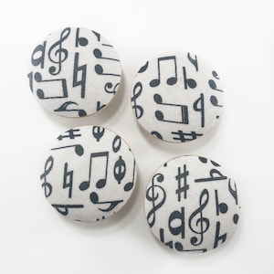 Puede incluir: Cuatro botones redondos cubiertos de tela. Cada botón presenta un fondo blanco con un patrón repetido de notas musicales y símbolos negros. Los botones tienen un borde de madera.