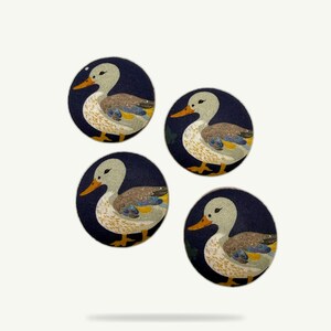 May include: Four round fabric patches with a duck illustration. Each patch features a duck with a light gray head, orange beak, and brown and yellow body, set against a dark blue background.