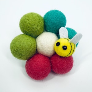 Pode incluir: Uma abelha de feltro repousa sobre um conjunto de bolas de feltro coloridas. As bolas são em tons de verde, branco e vermelho. A abelha é amarela com listras pretas e asas brancas. O fundo é branco.