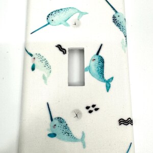 May include: White light switch cover featuring a pattern of teal narwhals with black horns. The narwhals are swimming on a white background with small black wave and dot designs. The cover has a rectangular opening.