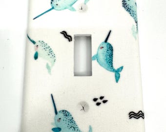 May include: White light switch cover featuring a pattern of teal narwhals with black horns. The narwhals are swimming on a white background with small black wave and dot designs. The cover has a rectangular opening.