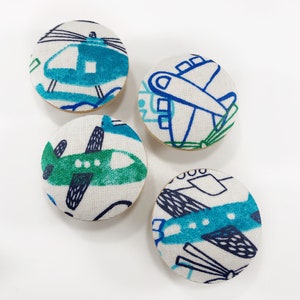 May include: Four round fabric buttons with a white background and blue, green, and black airplane and helicopter designs.