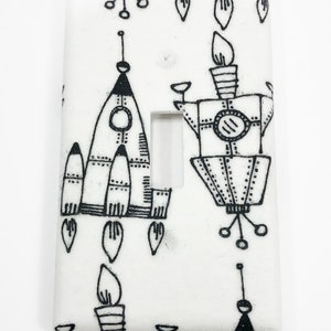 May include: A white light switch cover with a black and white pattern of rockets and spaceships. The pattern is repeated throughout the cover.
