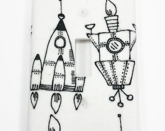 May include: A white light switch cover with a black and white pattern of rockets and spaceships. The pattern is repeated throughout the cover.