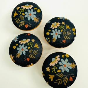 May include: Four round, dark navy blue fabric buttons with a floral pattern. The flowers are light blue, yellow, and pink, with green and orange leaves. The edges of the buttons are a light brown color.