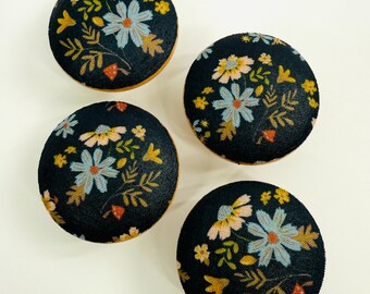 May include: Four round, dark navy blue fabric buttons with a floral pattern. The flowers are light blue, yellow, and pink, with green and orange leaves. The edges of the buttons are a light brown color.