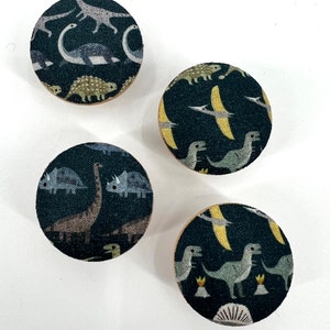 Pode incluir: Quatro botões redondos de madeira com fundo preto e um padrão de dinossauro colorido. Os botões têm um diâmetro de aproximadamente 5 cm.