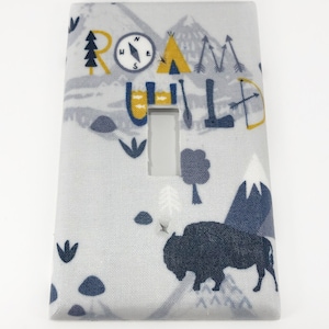 May include: A light gray light switch cover with a white background and a blue and yellow design. The design features a compass, a tent, a buffalo, and the words "Roam Wild".