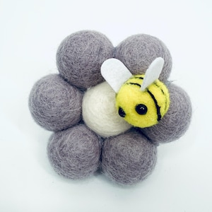 Może przedstawiać: Ręcznie wykonana filcowa pszczoła spoczywa na grupie szarych filcowych kulek, z jedną białą kulką pośrodku. Pszczoła jest żółta z czarnymi paskami i białymi skrzydłami. Kompozycja jest umieszczona na białym tle.