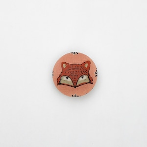 Pode incluir: Um botão redondo de tecido cor de pêssego com um design de rosto de raposa. A raposa tem uma pelagem marrom, olhos pretos e um nariz preto.