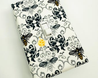 Puede incluir: Una cubierta decorativa para interruptor de luz con un fondo blanco adornado con un patrón de remolinos negros y grises, abejas y un panal amarillo. El interruptor es blanco.