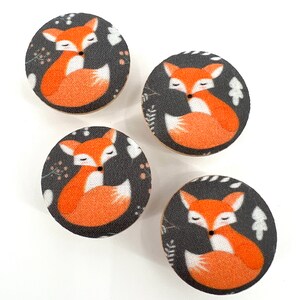 Könnte beinhalten: Vier runde Knöpfe mit schwarzem Hintergrund und einem weißen und orangefarbenen Fuchsdesign. Der Fuchs ist zusammengerollt, mit dem Schwanz um den Körper gewickelt. Die Knöpfe haben ein weißes und orangefarbenes Blumenmuster um den Fuchs herum.