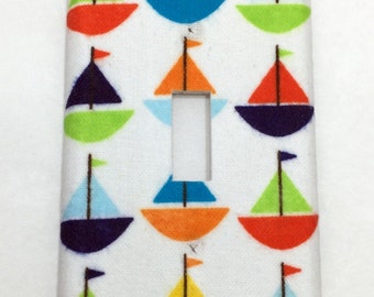 May include: A white light switch cover with a colorful sailboat pattern. The sailboats are red, orange, yellow, green, blue, and purple. The cover has a rectangular cutout for the light switch.