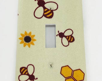 Puede incluir: Una tapa de interruptor de luz beige claro con un patrón de abejas, panales y girasoles. Las abejas son marrones con rayas amarillas y los panales son amarillos. Los girasoles son amarillos con centros marrones.