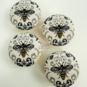 Peut inclure: Quatre boutons ronds recouverts de tissu avec un motif d'abeille. Chaque bouton présente une abeille noire et jaune sur un fond crème avec un motif damassé gris et noir. Les boutons mesurent environ 3,8 cm de diamètre.