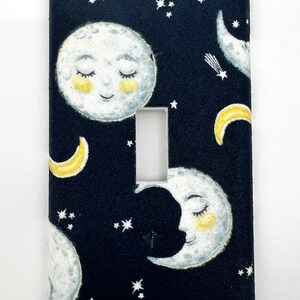 May include: A black light switch cover with a white and yellow moon and star pattern. The moon has a smiling face with closed eyes.