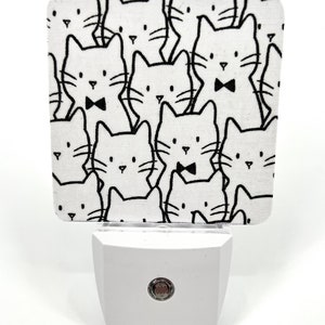 May include: A white night light with a black and white cat print cover. The night light has a round red button on the front.