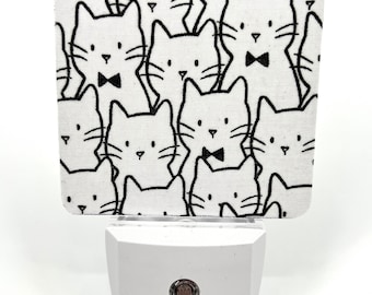 May include: A white night light with a black and white cat print cover. The night light has a round red button on the front.