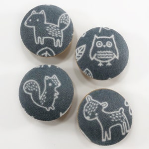 Op de afbeelding: Vier ronde, grijze stoffen knoppen met witte dierenontwerpen. De ontwerpen omvatten een vos, een uil, een eekhoorn en een hert.