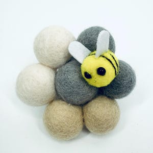 Pode incluir: Uma abelha de feltro amarela com listras pretas e asas brancas repousa sobre um grupo de bolas de feltro brancas, cinzentas e bege. Este conjunto artesanal é adequado para projetos de artesanato ou decoração de berçário.