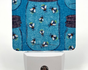 May include: A white night light with a blue fabric cover featuring a mason jar with fireflies inside. The fabric has a light blue background with a white, textured pattern.