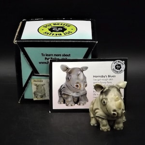 May include: A ceramic rhino figurine, "Hornsby's Blues," stands next to its box and a card. The rhino is gray with darker spots and a slightly open mouth. The box and card feature the "Pot Belly's" logo.