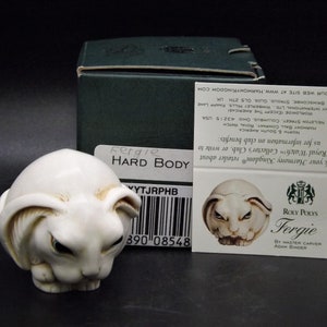 May include: A white ceramic figurine of a rabbit with black eyes and a closed mouth. The figurine is sitting on a white surface. The figurine is labeled "Fergie Hard Body" and is a Roly Poly's creation by master carver Adam Binder.