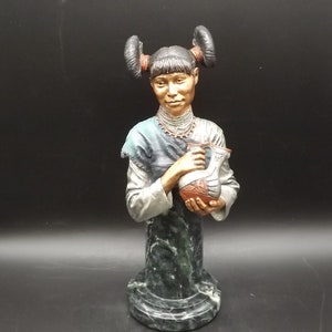 May include: A detailed bust of a woman with dark hair styled in two buns, wearing a blue and white top and holding a decorative vase. The base is green and gray, and the figure has a serene expression.