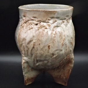May include: A handmade ceramic vase with a textured, organic design. The vase is off-white with brown accents and stands on three short legs. The vessel has a wide opening and a bulbous body, showcasing a unique, rustic aesthetic.