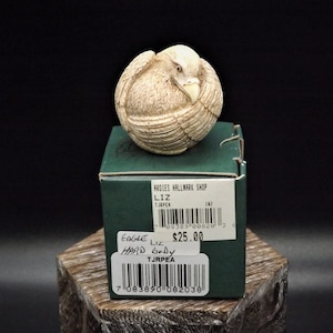 May include: A white ceramic eagle figurine with its wings wrapped around its body, sitting on a green box with the text "ARDIES HALLMARK SHOP", "LIZ", "TJRPER", "182", "EAGLE", "LIZ", "HARD BODY", "TJRPEA", "$25.00", and barcodes.