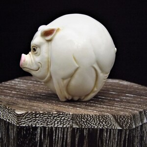 May include: A white ceramic sculpture of a pig, with a pink snout and ears, is sitting on a wooden base.