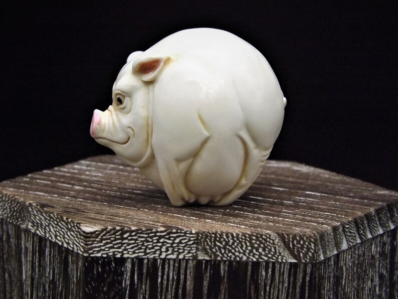 May include: A white ceramic sculpture of a pig, with a pink snout and ears, is sitting on a wooden base.