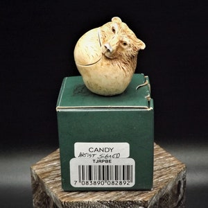 May include: A small ceramic bear figurine in a curled up position. The bear is brown and white and has a small black nose. The figurine is sitting on a green box with the words "CANDY ARTIST SIGNED TJARPE" and a barcode.