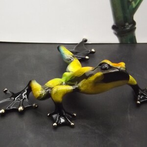 May include: A detailed sculpture of a frog in a dynamic pose. The frog features a vibrant color scheme with yellow, black, and green hues. Its feet are black with silver accents. The frog is positioned on a dark surface.