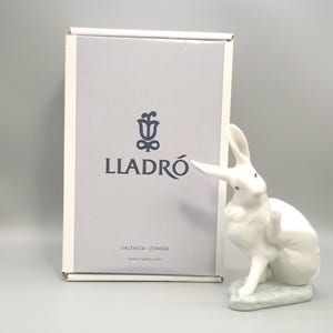 May include: A white porcelain rabbit figurine in a seated position, scratching its ear, placed next to a rectangular white box featuring the dark gray Lladro logo, a stylized flower icon, and the text Valencia España and www.lladro.com.