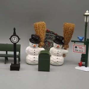 May include: A set of miniature Christmas village decorations, including two snowmen holding brooms, a green mailbox, a green bench, a clock tower, and a street lamp with a "Village Realty FOR SALE" sign.