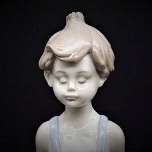 May include: A porcelain figurine of a young boy with closed eyes, wearing a blue and white outfit. The figurine is made of white porcelain with a light brown hair color.