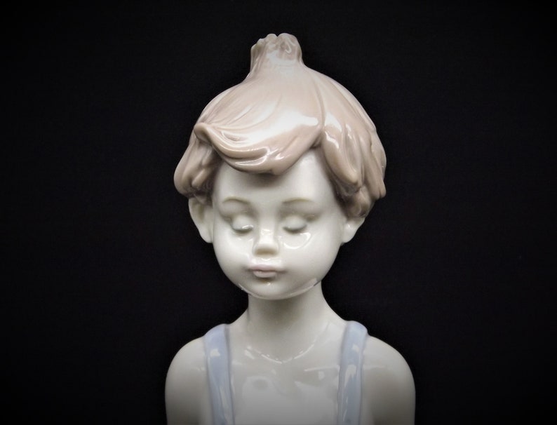 May include: A porcelain figurine of a young boy with closed eyes, wearing a blue and white outfit. The figurine is made of white porcelain with a light brown hair color.