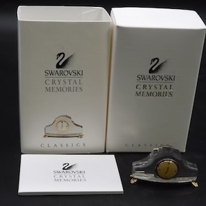 May include: A Swarovski Crystal Memories clock figurine with a gold-tone clock face and gold-tone feet. The clock is displayed with its original packaging, including a white box and a small card, all bearing the Swarovski logo.