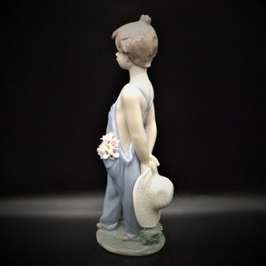 May include: A porcelain figurine of a young boy wearing blue overalls and holding a straw hat. He has brown hair and is holding a bouquet of flowers in his pocket.