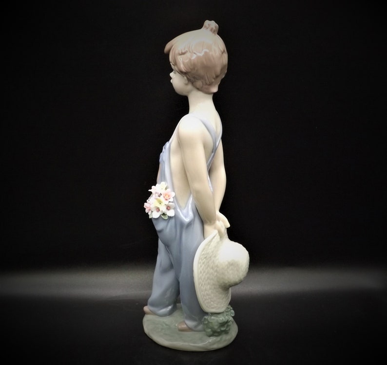 May include: A porcelain figurine of a young boy wearing blue overalls and holding a straw hat. He has brown hair and is holding a bouquet of flowers in his pocket.