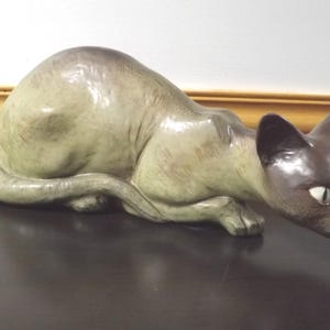 May include: A ceramic cat figurine in a crouching position with a light beige body, a dark brown face, and dark brown ears. The cat has white almond-shaped eyes and a smooth, glossy finish, depicted in a low-profile, alert posture.