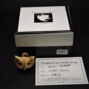 May include: An owl mask figurine with a certificate of authenticity. The mask is light brown with large eyes and a curved beak. The box is white and black with the text "Adam Binder Editions". The certificate reads "Owl Mask" and "ABE Netsuke's".