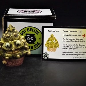 May include: A quirky, green "Green Gleamer" Christmas tree ornament with a smiling face, presented with a box and a card detailing the history of Christmas trees. The card includes the text "Seasonals" and "Green Gleamer". The figurine is set on a decorative base.